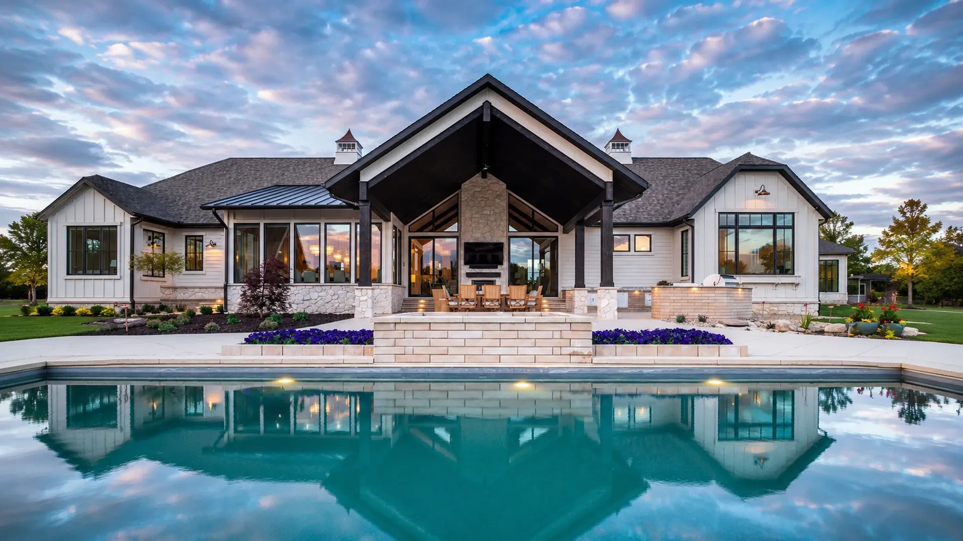 Luxury custom inground pool and modern farmhouse outdoor living design at 37291 Seabiscuit Park Pl in Middleburg VA, featuring a reflective geometric swimming pool, premium stone patio, covered outdoor lounge, stone fireplace wall, outdoor kitchen and grill area, large glass doors, architectural lighting, flower beds, natural stone accents, and resort-style backyard construction by AM Developers DMV. This project highlights luxury custom pool construction, gunite pool builders, shotcrete swimming pool craftsmanship, high-end patios and outdoor living spaces, pool decking materials, natural stone coping, landscape lighting for pools, outdoor kitchen and living, and premium hardscape design in Northern Virginia.